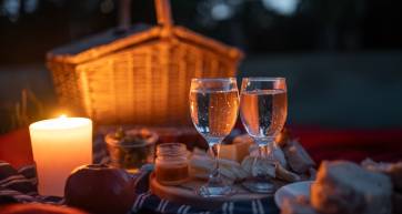 Two glasses of sparkling wine by candle and basket, surrounded by delightful spread of bread, fruit, and cheese, all set for intimate picnic as dusk falls in peaceful outdoor location.