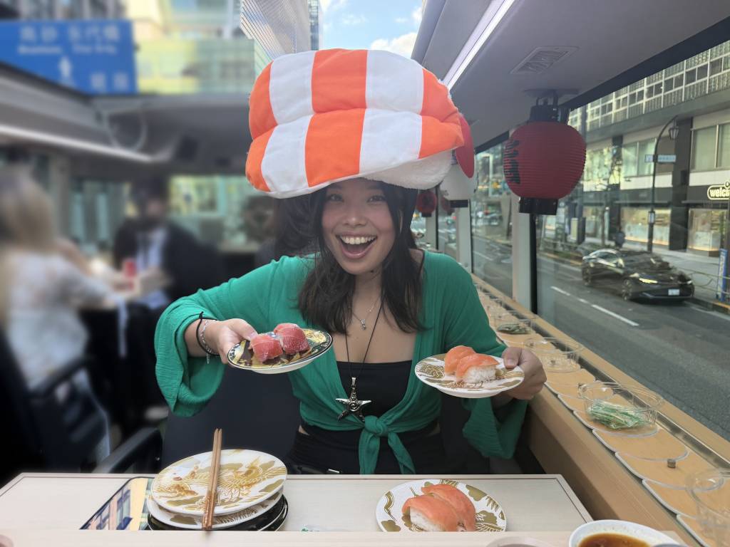 Woman in a sushi hat holding up plates of tuna and salmon nigiri on the open top deck of the Sushi Bus in Tokyo