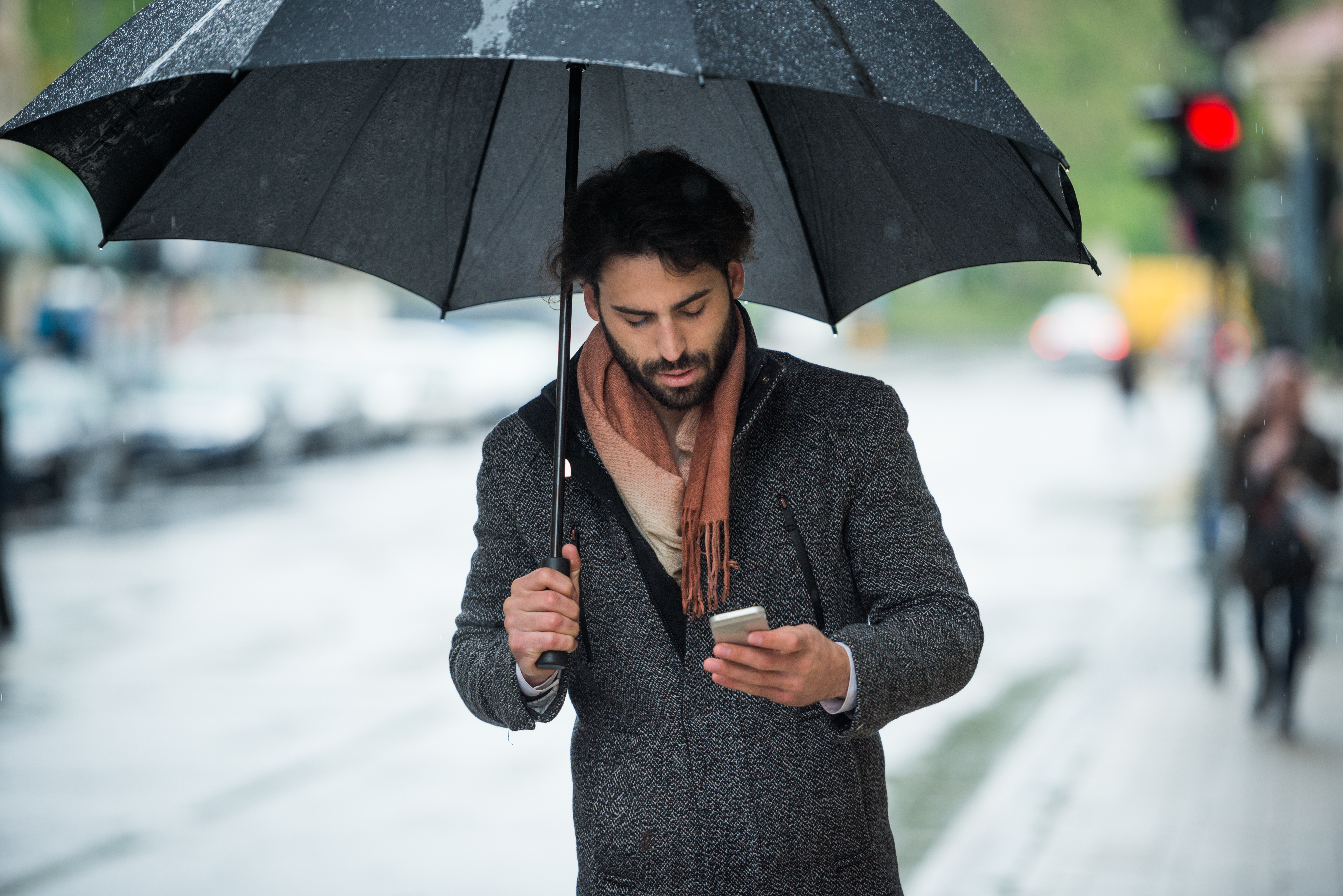 Young man walking in the city street with umbrella on a rainy day in October, and looking at his mobile phone. Crossroad and traffic light can be seen in the background.