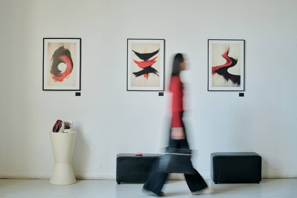 Individual with blurred figure walking past modern art pieces in gallery creating sense of motion, black and red abstract paintings displayed on white wall